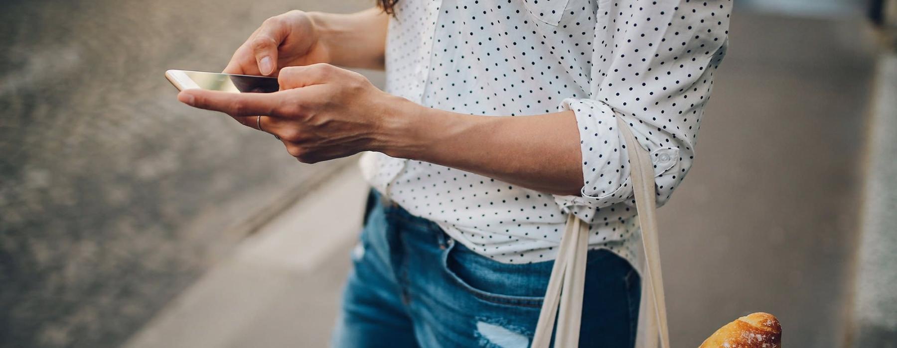 woman walks down the street and texts with a bag of groceries on her arm