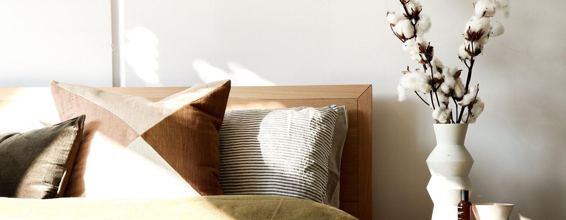 swath of sunlight shines across a well made bed and bedside table with a vase of cotton plants